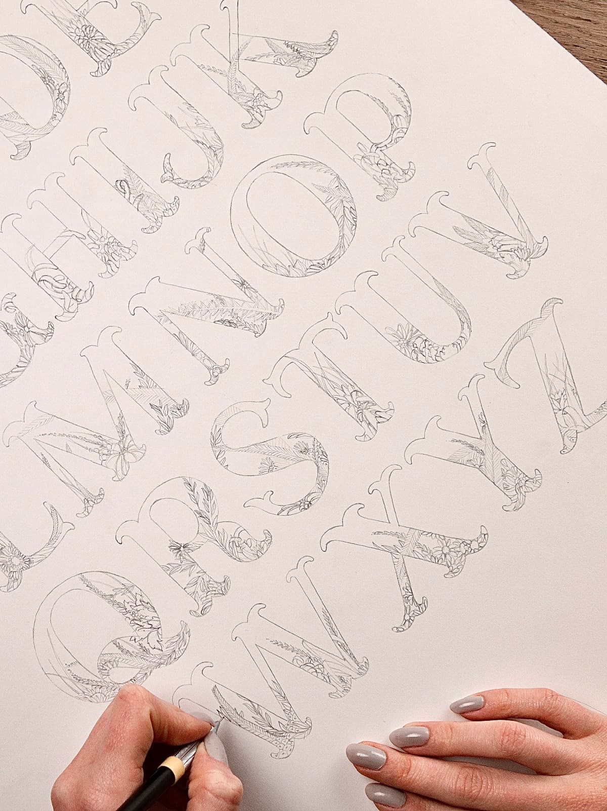 Close-up of hands using a pencil to sketch intricate floral details onto a paper alphabet letterform.